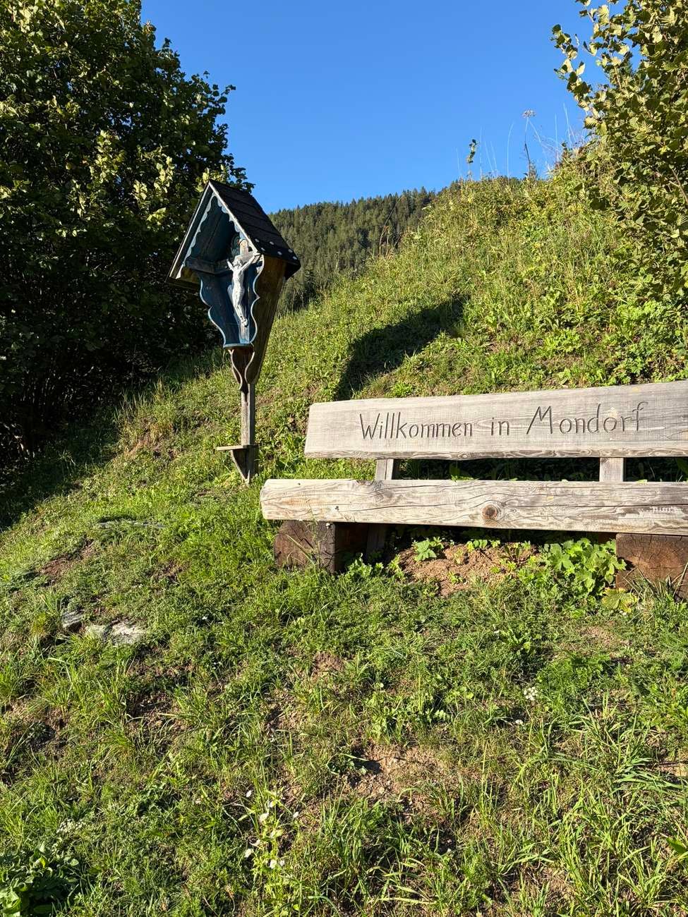 Holzbank „Willkommen in Mondorf“ mit Wegkreuz am Wiesenhang